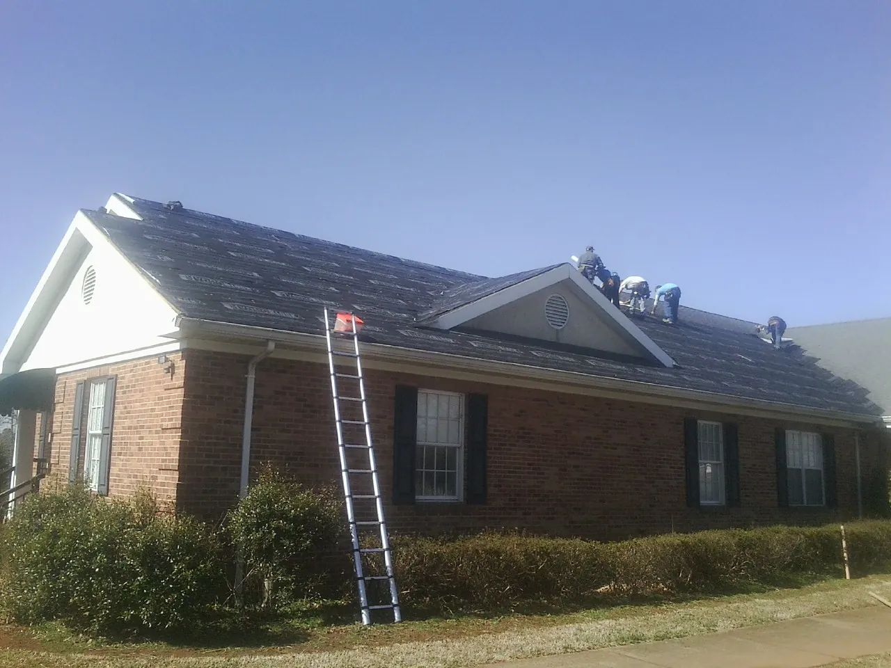 Crew tearing shingles off a brick ranch roof with a ladder set — Emergency Roof Repair work by our crew in Hill
