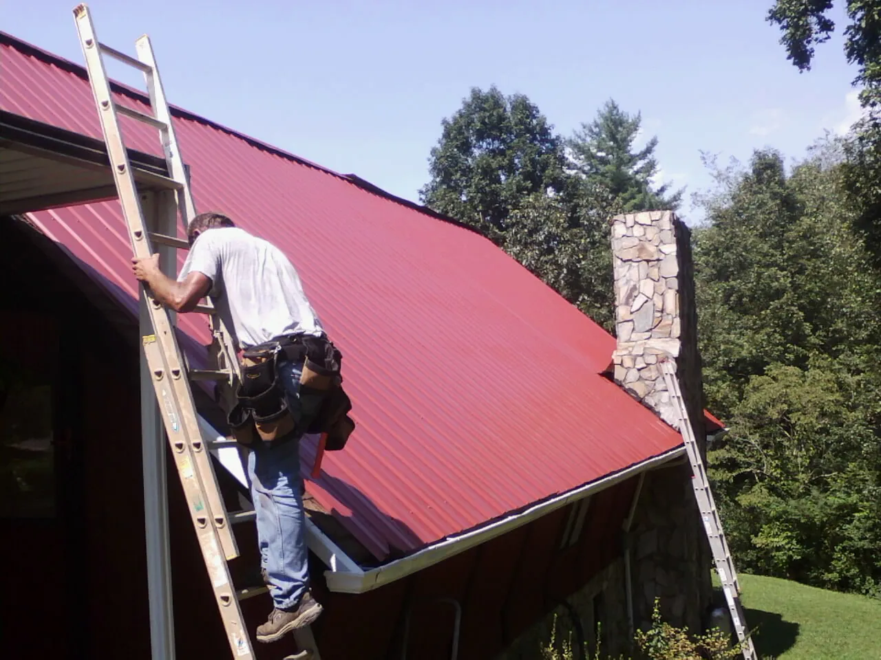 Roofer on a ladder beside a red metal roof and stone chimney — Soffit & Fascia Repair by Roofing Brothers in Hill